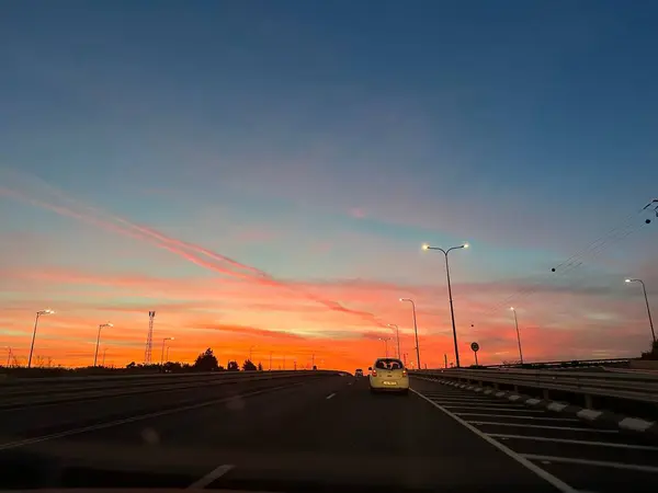 Gün batımında gökyüzünde canlı turuncu ve pembe renkleri olan bir otoyolun manzarası. Sokak lambaları yolu aydınlatıyor ve ileride bir arabanın sürdüğü görülüyor..
