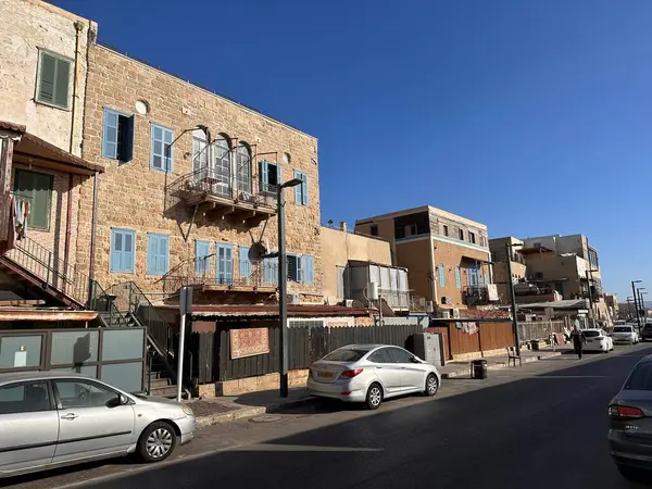 Taş cepheli ve mavi pencereli tarihi binaların sokak manzarası. Sahnede yol boyunca park etmiş arabalar, açık mavi gökyüzü altında eski mimari ve modern araçların bir karışımını sergiliyor..