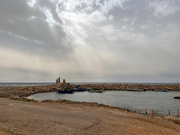 Kayalık oluşumlar ve bulutlu gökyüzü ile çevrili küçük bir liman içeren sakin bir sahil manzarası. Güneş ışığı bulutların arasından doğar, suyu aydınlatır..