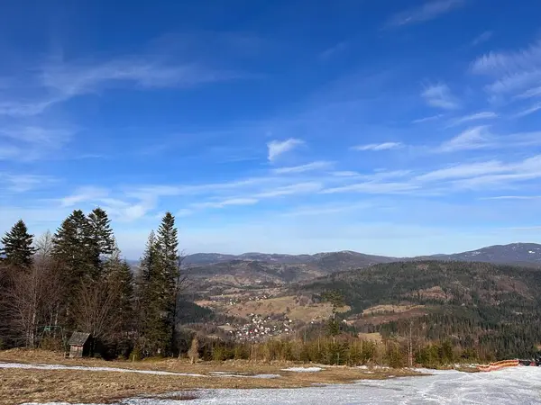 Berrak mavi gökyüzünün altındaki dağlık bir manzaranın manzarası. Ön planda kar parçaları olan çimenli bir alan yer alırken, arka planda kayan tepeler ve vadide yuva yapmış küçük bir köy yer alıyor..