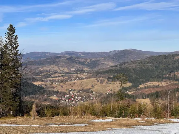 Yuvarlanan tepelerin manzarası ve açık mavi gökyüzünün altında ağaçlar ve dağlarla çevrili bir vadide yuva yapmış küçük bir köy. Yerde kar parçaları var, bu da kışı gösteriyor..
