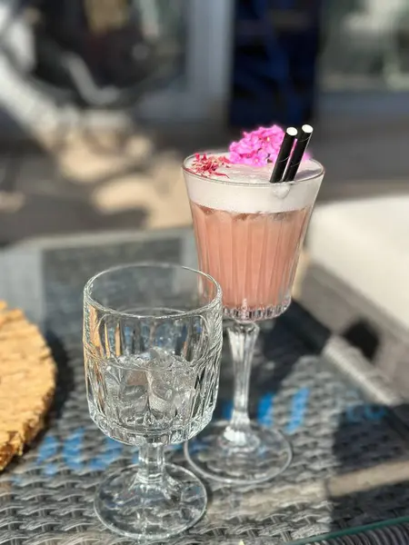 Uzun bir bardakta köpüklü ve çiçekli pembe bir kokteyl. Hasır bir masada boş bir bardağın yanında..