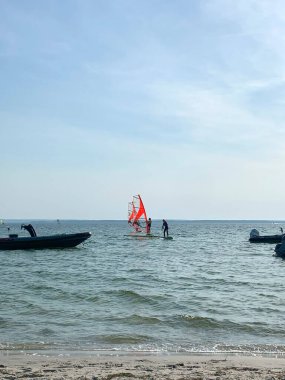 Rüzgâr sörfü tahtasında parlak kırmızı yelkenli iki kişinin olduğu sakin bir sahil sahnesi. Arka planda, küçük tekneler berrak mavi bir gökyüzünün altında durgun sularda demirliyorlar..