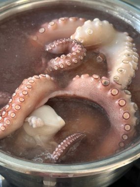 Ahtapotların, dokunaçlarını ve dokularını sergiledikleri bir tencere suya batırılmış yakın plan görüntüsü. Su biraz bulanık görünüyor, bu da yemek hazırlığını gösteriyor..