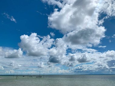 Sakin bir okyanus üzerinde tüylü beyaz bulutlarla dolu engin bir gökyüzü içeren sakin bir deniz manzarası. Küçük tekneler uzak bir yere demir atarak huzurlu bir atmosfer yaratıyor..