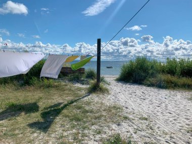 Renkli çamaşırların asılı olduğu sakin bir sahil sahnesi kumlu bir yola çıkıyor. Gökyüzü kabarık bulutlarla aydınlık ve arka planda sakin bir deniz görülebilir..