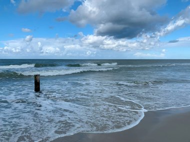 Sahile doğru hafif dalgaların dalgalandığı sakin bir sahil manzarası, tek bir yıpranmış direğin kumsalda durduğu. Gökyüzü parçalı bulutlu, mavi ve beyaz bulutların karışımını sergiliyor.
