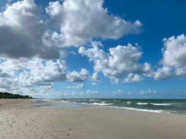 Yumuşak dalgaların kıyıya vurduğu, tüylü beyaz bulutlarla dolu, parlak mavi bir gökyüzünün altında sakin bir sahil sahnesi. Kumsalın çoğu boş, huzur verici..