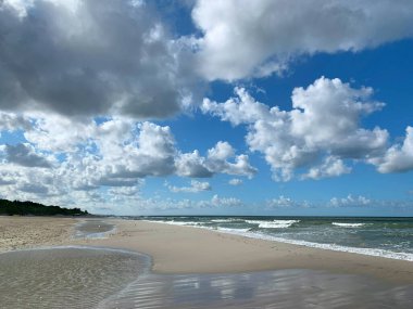 Yumuşak dalgaların kıyıya vurduğu tüylü beyaz bulutlarla dolu sakin bir sahil sahnesi. Kumlu sahil mesafeye kadar uzanıyor ve sakin bir atmosfer yaratıyor..