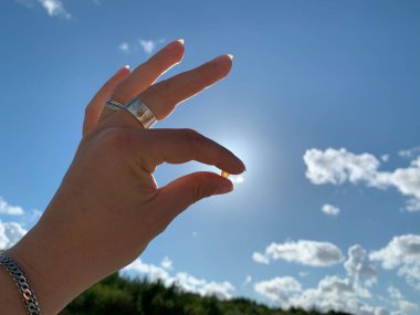 Küçük bir nesneyi bulutlu açık mavi bir gökyüzüne karşı tutan bir el siluet etkisi yaratıyor. Güneş, ışık ve gölge arasındaki zıtlığı vurgulayarak el tarafından kısmen gizleniyor..