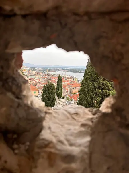 Kırmızı çatılı binalar, ağaçlar ve arka planda bir su kütlesi olan taştan bir duvarın içinden bir sahil şehri manzarası..