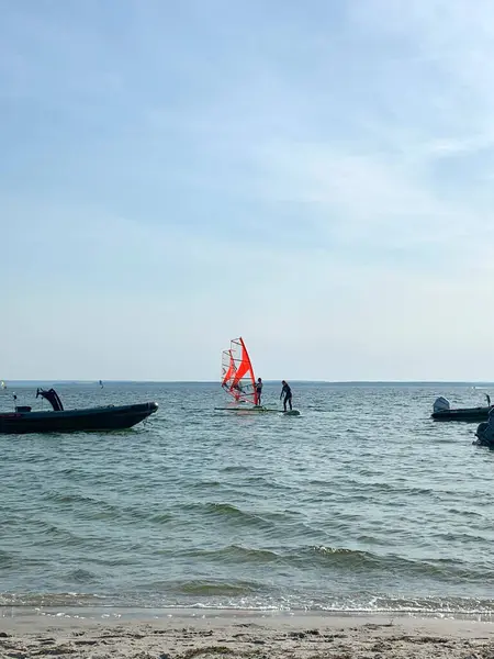 Rüzgâr sörfü tahtasında parlak kırmızı yelkenli iki kişinin olduğu sakin bir sahil sahnesi. Arka planda, küçük tekneler berrak mavi bir gökyüzünün altında durgun sularda demirliyorlar..