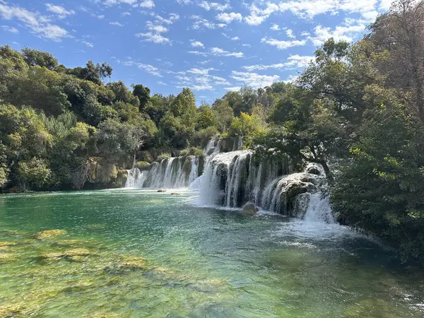 Manzaralı şelale, bulutlu bir gökyüzünün altında yemyeşil bir havuzun içinde berrak bir turkuaz havuza dökülüyor..