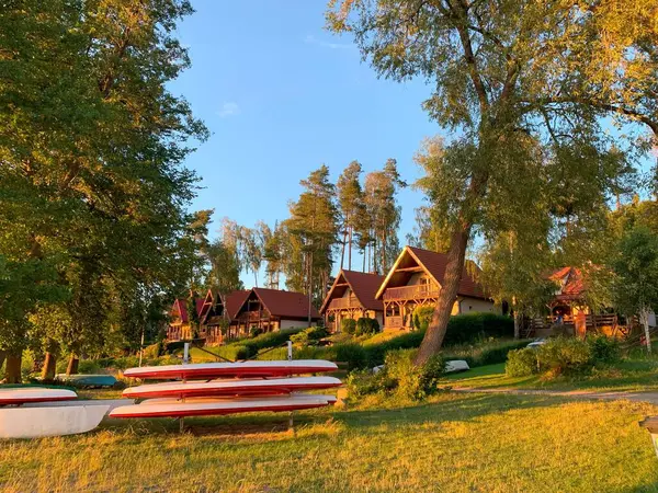 Bereketli yeşillikler ve uzun ağaçlarla çevrili ahşap kulübelerin bulunduğu sakin bir göl manzarası. Sıcak güneş ışığı manzaranın üzerine altın bir renk saçar ve sakin atmosferi güçlendirir..