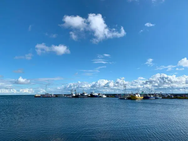 Bir kaç balıkçı teknesinin bulunduğu sakin bir liman sahnesi berrak mavi bir gökyüzünün altında, tüylü beyaz bulutlarla kenetlenmiş. Ortam sakin ve davetkar. Denizcilik teması için ideal..