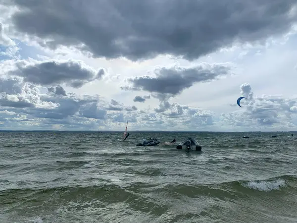 Bulutlu bir gökyüzünün altında dalgalı bir denizin manzarası. Uzakta kayıklar ve uçurtma sörfçüleri var. Dalgalar dalgalı, dinamik bir atmosfer yaratıyor..