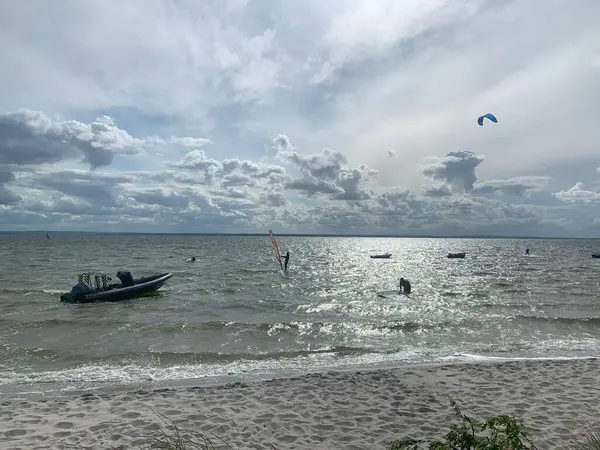 Sakin bir sahil sahnesi. İçinde küçük tekneler ve rüzgar sörfçüleri olan sakin bir deniz. Gökyüzü parçalı bulutludur. Güneş ışığını su yüzeyine yansıtarak sakin bir atmosfer yaratır..