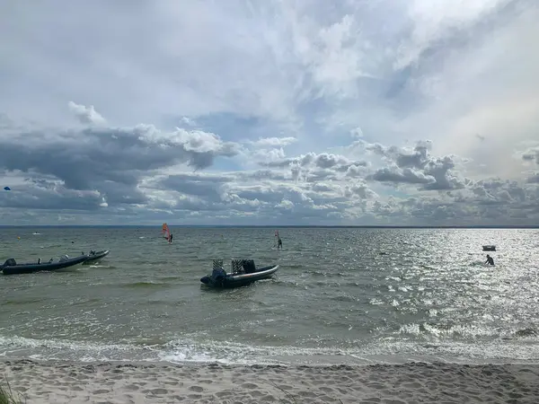 Bulutlu bir gökyüzünün altında sakin sularda kayıkların olduğu sakin bir sahil sahnesi. Güneş suya yansıyarak, parıldayan bir etki yaratıyor. Rüzgâr sörfçüleri uzaktan görülebiliyor ve sakin ortama etkinlik katıyor.