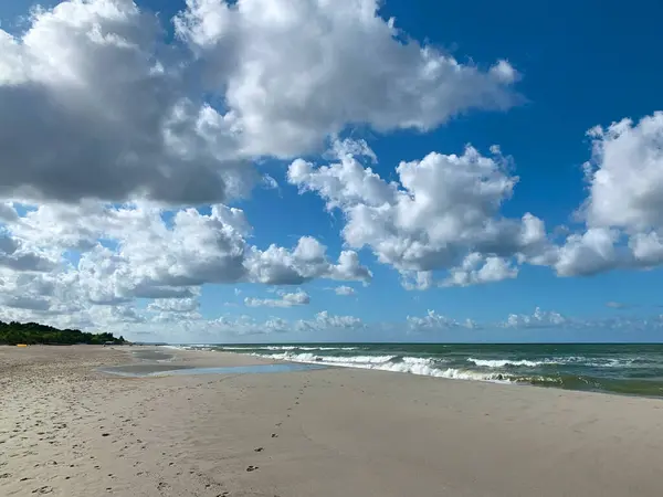 Yumuşak dalgaların pofuduk beyaz bulutlarla dolu açık mavi bir gökyüzünün altında kıyıya vurduğu sakin bir sahil sahnesi. Kumsalın büyük bölümü boş. Rahatlama ve sükûnet dolu..