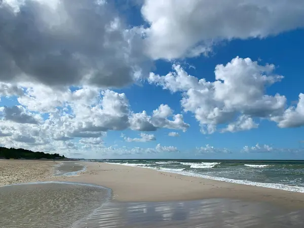 Yumuşak dalgaların kıyıya vurduğu tüylü beyaz bulutlarla dolu sakin bir sahil sahnesi. Kumlu sahil mesafeye kadar uzanıyor ve sakin bir atmosfer yaratıyor..