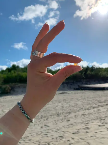 Kumlu bir sahilde, tüylü bulutlu parlak mavi bir gökyüzüne karşı küçük bir nesneyi tutan bir el. El, yüzüklerle ve bileziklerle süslenir. Rahat bir plaj havası katar..