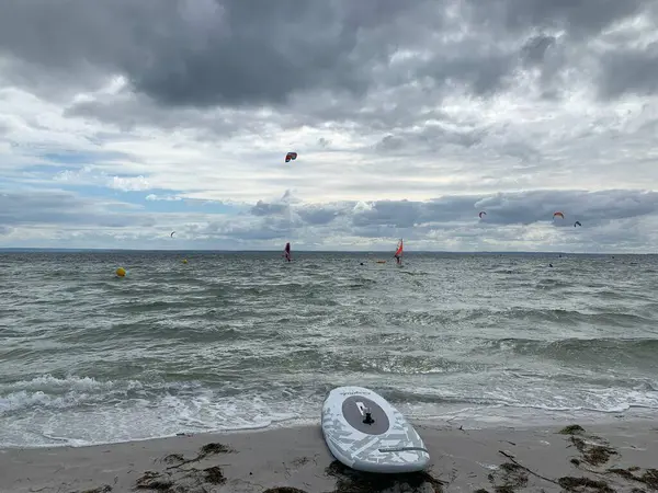 Sahilde, bulutlu bir gökyüzüne karşı uçurtma sörfçülerinin olduğu sakin bir plaj sahnesi. Dalgalar yavaşça kumlu sahilde dolaşarak sakin bir atmosfer yaratıyor..