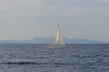 Sakin bir denizde süzülen yalnız bir yelkenli, ufukta görünen uzak dağlar bulutlu bir gökyüzünün altında..