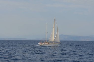 Sakin mavi sularda süzülen bir yat beyaz yelkenli ile, uzak dağlar ve berrak bir gökyüzü karşı.