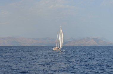 Sakin bir denizde süzülen yalnız bir yelkenli. Arka planda uzak dağlar, berrak bir gökyüzünün altında..