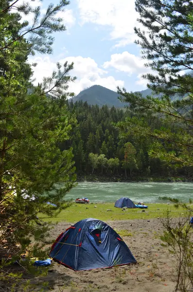 Nehir kenarındaki bir kamp alanının manzarası, kumlu zemine kurulmuş mavi çadırlar yemyeşil ağaçlar ve dağlarla çevrili arkaplanda parçalı bulutlu bir gökyüzünün altında..