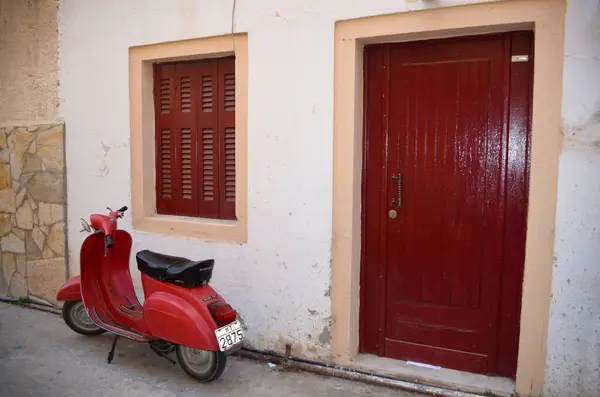 Kırsal bir kapının yanında park etmiş, ahşap panjurlu klasik kırmızı bir scooter. Duvar desenli ve açık renkli. Sahneye cazibe katıyor..