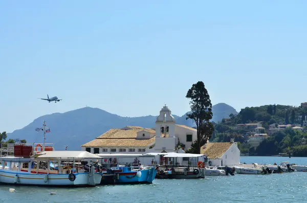 Ön planda tekneler, çan kulesi olan büyüleyici beyaz bir kilise ve açık mavi gökyüzüne karşı uçan bir uçak..