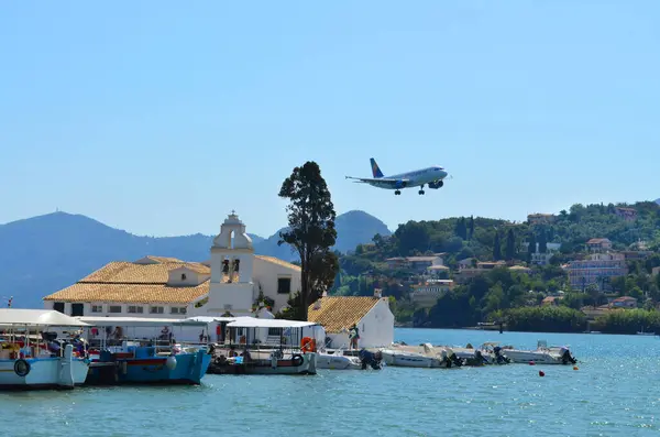 Tekneli küçük bir liman, büyüleyici beyaz bir kilise ve açık mavi gökyüzüne karşı suyun üzerinde alçaktan uçan bir uçağın yer aldığı pitoresk bir manzara..