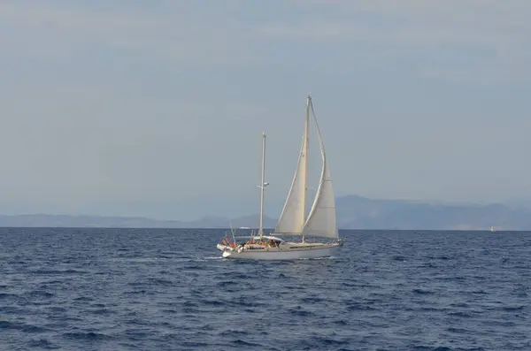 Sakin mavi sularda süzülen bir yat beyaz yelkenli ile, uzak dağlar ve berrak bir gökyüzü karşı.