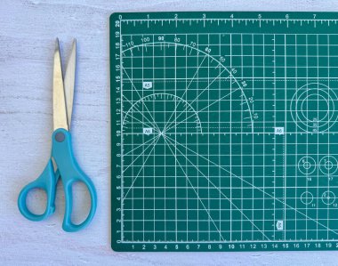 Green self-healing cutting mat with measurement grid beside turquoise craft scissors on light wooden surface. Perfect setup for DIY, paper crafts, sewing, scrapbooking, and other handmade projects.