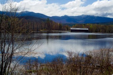 Gorham NH, Başkanlık sıradağları ve gökyüzünün bulutlu yansımaları. Uzakta kırmızı kaplı bir köprü.