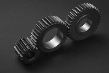 Üç siyah dişli koyu gri bir yüzey üzerinde birbirine yakın konumdadır. Dişlilerin pürüzsüz, metalik dişleri ve merkezi delik var. Aydınlatma dramatik, derin gölgeler bırakıyor.