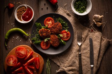 Koyu renkli, karamsar bir fotoğrafta siyah bir tabakta servis edilen bir tabak falafel taze otlar, domatesler ve sosla süslenmiş. Tabak kırsal bir ahşap masada duruyor.