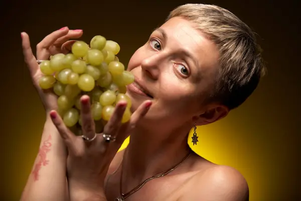 Kısa sarı saçlı genç bir kadın büyük bir demet yeşil üzümü yüzünün önünde tutuyor, hafifçe şaşırmış ve neşeli görünüyor. O, gümüş bir kolye ve sağ elinde bir yüzük takıyor. Sol kolunda bir ejderha dövmesi var..