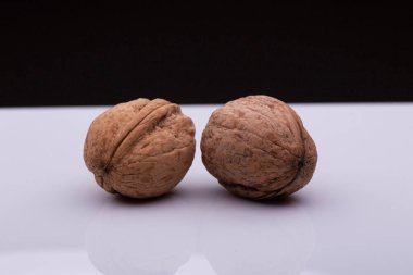 Beyaz bir yüzeyde yan yana duran iki ceviz ayna yansıması yaratır. Cevizlerin kabukları pürüzlü, desenli ve görünen çatlakları var. Arka plan karanlık.