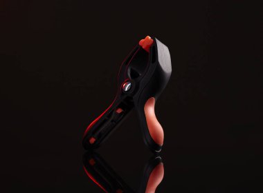 Siyah ve kırmızı bir kıskaç yansıtıcı siyah bir yüzeye dayanır. Klempin sapında kırmızı aksanlı, parlak ve modern bir tasarımı var. Işıklandırma çarpıcı, yüksek kontrastlı bir etki yaratıyor.