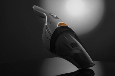 Pürüzsüz, siyah ve gri bir elektrikli süpürge belirgin bir şekilde koyu, yansıtıcı bir arka plana karşı sergilenir. Parlak, dairesel bir ışık kaynağı doğrudan vakumun üzerinde parlar.