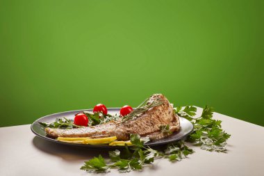 Büyük, ızgara balık bifteği taze biberiye filizleri, maydanoz ve birkaç canlı kırmızı vişne domatesiyle süslenmiş koyu gri bir tabakta dinlenir. Biftek açık bej bir yüzeyde servis edilir.