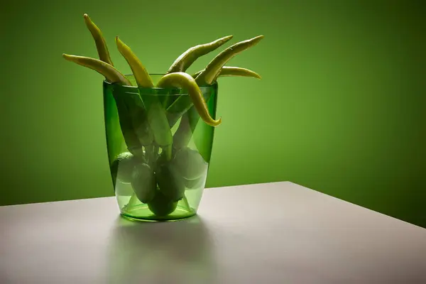 Canlı bir yeşil biber demeti berrak bir cam vazonun içinde düzenlenir. Vazo açık gri bir masanın üzerinde duruyor ve arka plan olarak som yeşil bir duvar kullanılıyor. Işıklandırma dramatik.