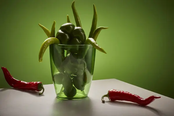 Yeşil biberle dolu cam bir vazo beyaz bir masanın üzerinde duruyor. Vazonun yanındaki masada tek bir kırmızı kırmızı biber duruyor. Arka plan koyu yeşil bir renktir. Işıklandırma dramatik.