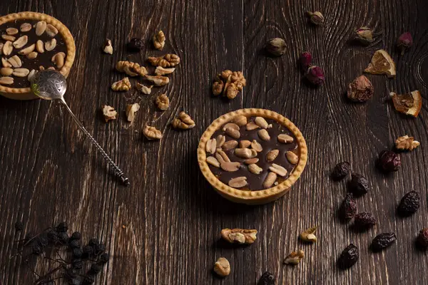 İki çikolatalı turta, dağılmış cevizler, kurutulmuş meyveler ve gül yapraklarıyla çevrili, koyu ahşap bir yüzeyde otururlar. Turtalardan birinin yanında gümüş bir kaşık duruyor. Turtaların altın-kahverengi bir kabuğu var.