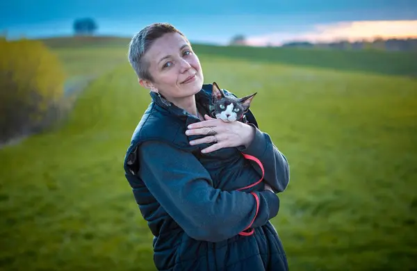 Kanadalı bir Sphynx kedisini kollarında tutarken dışarıda yürüyen bir kadını gösteren samimi bir fotoğraf. Görüntü, insan ve evcil hayvan arasındaki benzersiz bağı vurgular, doğal ışıkla.