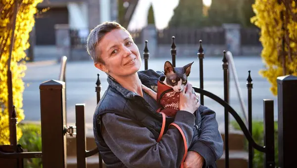 Kanadalı bir Sphynx kedisini kollarında tutarken dışarıda yürüyen bir kadını gösteren samimi bir fotoğraf. Görüntü, insan ve evcil hayvan arasındaki benzersiz bağı vurgular, doğal ışıkla.