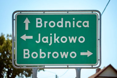 Polonya 'da güneşli bir günde sürücülere rehberlik eden Brodnica, Jajkowo ve Bobrowo' yu gösteren beyaz yazı ve oklarla yeşil yönlü yol işareti.