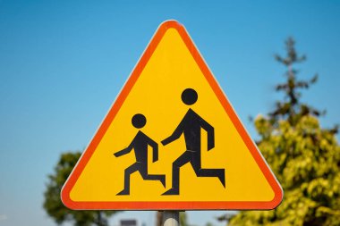 Karşıya geçen çocuklar için üçgen şeklinde bir uyarı işareti. Açık mavi gökyüzüne karşı çekilen fotoğraflar, sürücülere okul ve yaya alanlarının yakınında yavaşlamalarını hatırlatıyor..